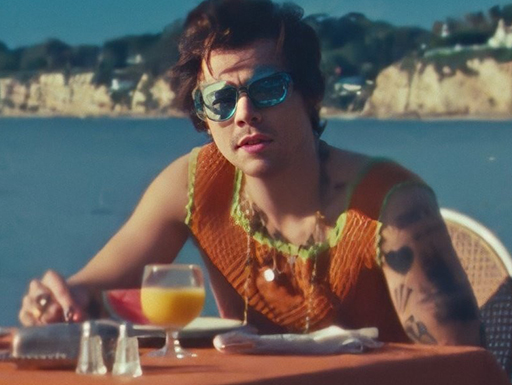 Harry Styles, de óculos escuros, sentado em uma mesa na beira da praia no clipe de "Watermelon Sugar"