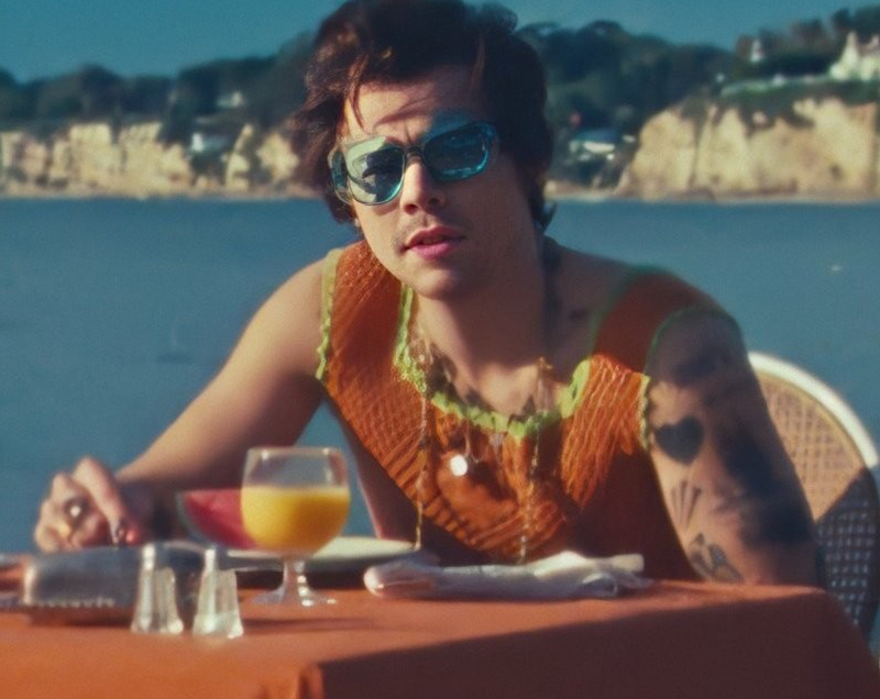 Harry Styles, de óculos escuros, sentado em uma mesa na beira da praia no clipe de "Watermelon Sugar"