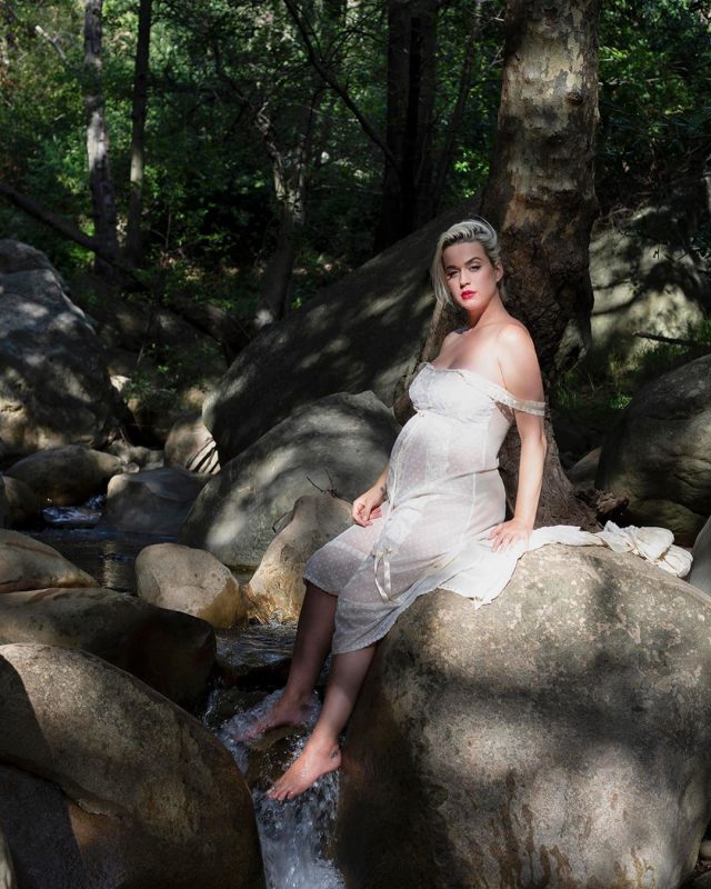 Katy Perry aparece na beira de um córrego, sentada em uma rocha, vestida de branco e grávida
